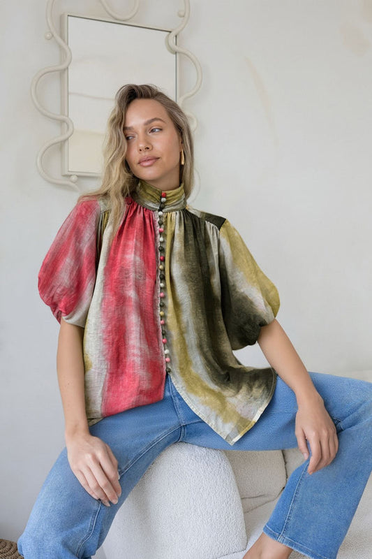 Woman wearing a colorful blouse with puffed sleeves and blue jeans sitting on a white chair.