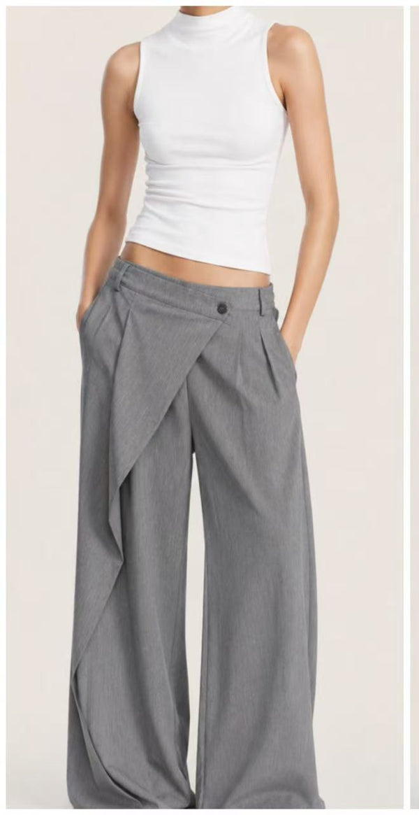 A model wearing a white ribbed mock-neck tank top and high-waisted, wide-leg gray wrap pants with side pleats and a button closure, set against a plain light beige background.