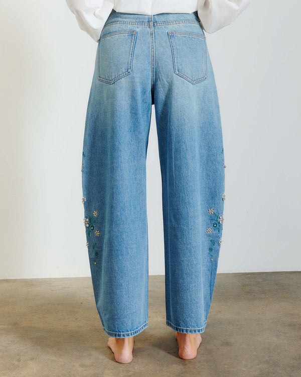 Rear view of a person wearing wide-leg light blue denim jeans with floral embroidery down the outer sides, paired with a white top against a plain indoor wall.