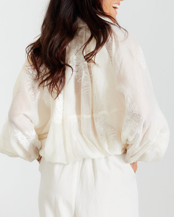 Back view of a woman wearing a cream semi-sheer embroidered batwing jacket with balloon sleeves, paired with matching wide-leg trousers.
