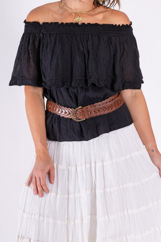 Woman wearing a black off-shoulder cotton top with a brown belt and white pants on a white background